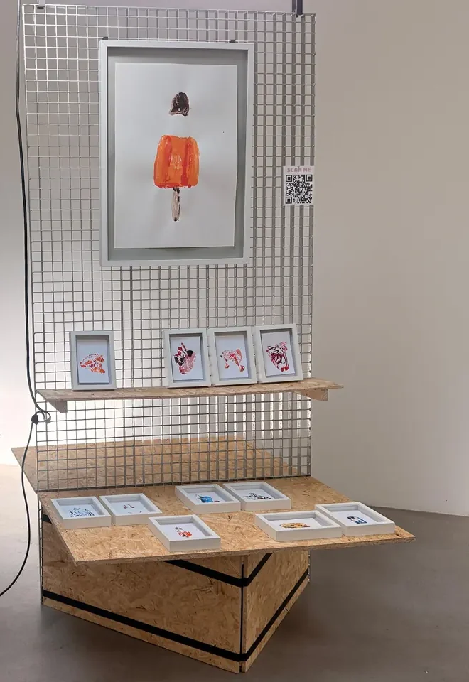 Testaufbau: an einer Gitterwand hängt ein gerahmtes Werk, daneben ein QR-Code. Darunter ein Regalbrett mit vier kleinen Rahmen, davor ein Möbel aus Spanplatten, auf dem mehrere kleine Arbeiten flach ausliegen.