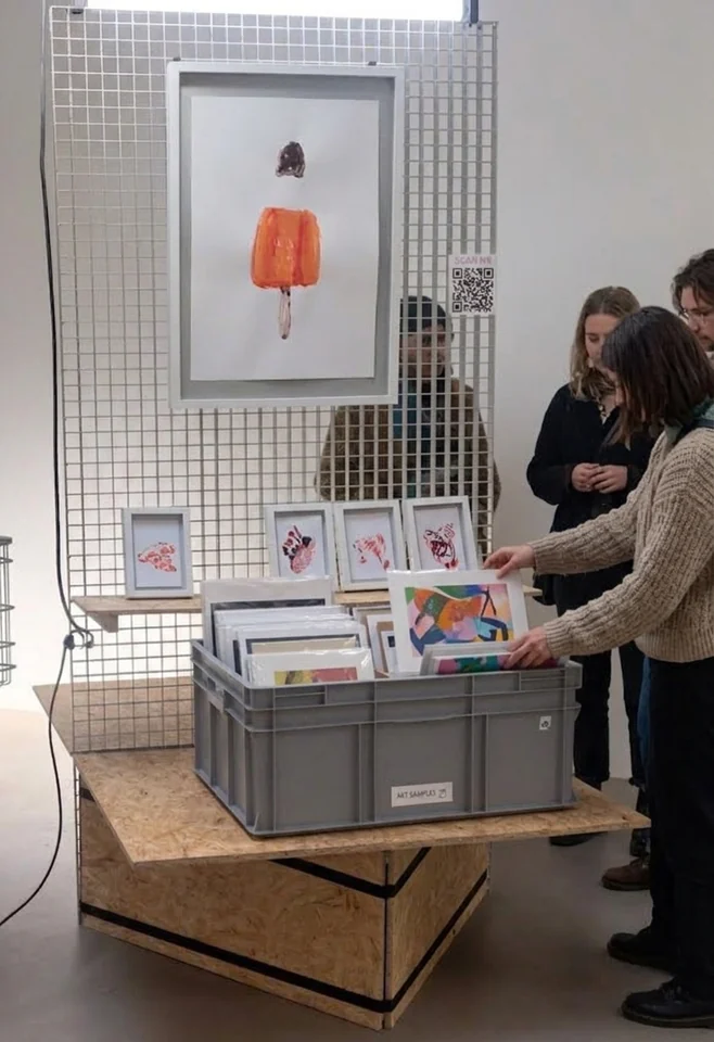 Auf dem Spanplattenmöbel steht eine graue Box mit der Aufschrift „Art Samples“, darin stehen gerahmte Arbeiten aufrecht nebeneinander. Eine Besucherin zieht eine davon heraus, weitere Leute stehen daneben.